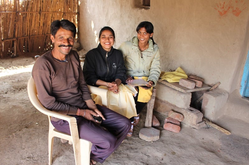 A man and two women sit inside a house in India, smiling at the camera.