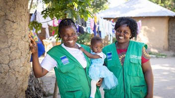Two women in ChildFund vests in Kenya stand holding a little girl wearing a tutu, smiling.