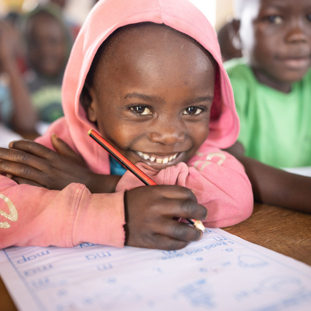 A child in Kenya sits at a desk in his classroom, studying and smiling at the camera beneath his hoodie.