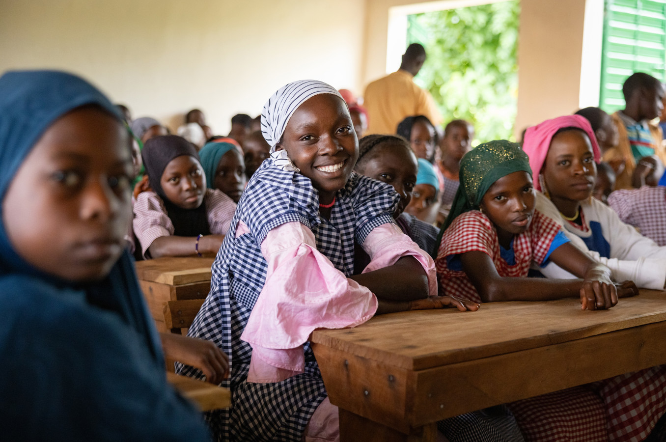 A girl in Guinea wearing a colorful head covering sits in a crowded classroom at a wooden desk, smiling at the camera.