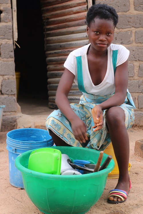 Teenage girl in Zambia washes dishes while looking at camera.