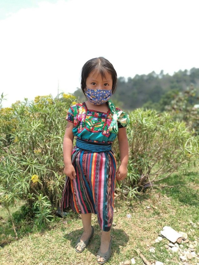 Small girl in Guatemala stands outside wearing mask looking at the camera.